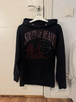 Svart hoodie South of Heaven - Svart hoodie med huva och magficka. Framsidan har ett stort tryck med texten 'South of Heaven' i rött och en grafisk design med lejon och sikte. Perfekt för dig som gillar streetwear och vill sticka ut.