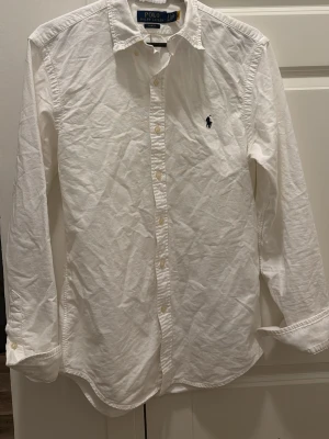 Vit skjorta  Polo Ralph Lauren - Vit skjorta från Polo Ralph Lauren i slim fit-modell. Skjortan har långa ärmar, knappar framtill och den ikoniska broderade loggan på bröstet. Tillverkad i mjuk bomull och passar perfekt till en clean och stilren look.