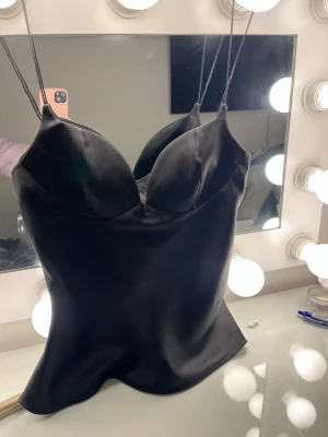 Svart satin korsettopp med tunna band - Supersnygg svart korsettopp i satin med tunna axelband och formade kupor. Toppen har en figurnära passform och en lätt glansig yta som ger en lyxig känsla. Perfekt att styla med jeans eller kjol för en trendig look.