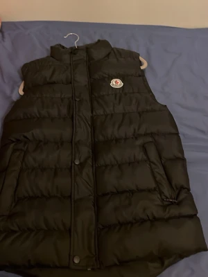 Svart dunväst från Moncler  - Snygg svart dunväst från Moncler med pris kan diskuteras quiltad design och Moncler-logga på bröstet. Västen har hög krage, dragkedja och tryckknappar framtill samt två snedställda fickor. Perfekt för lager-på-lager och streetwear. 