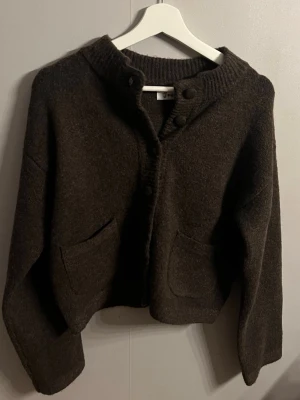 Mörkbrun ullkofta med fickor - Säljer en mörkbrun,  kofta i ull med två stora fickor framtill och ribbad krage. Koftan har knäppning med stora knappar och en avslappnad passform. Perfekt för lager på lager och en chill look.