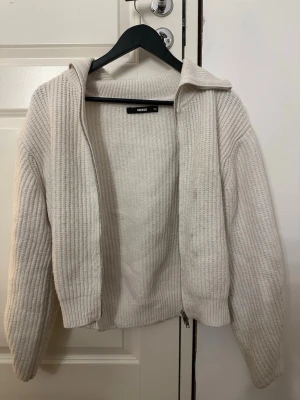 Beige ribbad ziptröja från Bik Bok - Säljer en vit ribbstickad ziptröja/ kofta från Bik Bok. Tröjan har hög krage, långa ärmar och dragkedja framtill. Materialet är mjukt och tjockt, perfekt för lager på lager. Enkel och stilren design som passar till det mesta. Säljer för billigt pga defekten på högra sidan😊