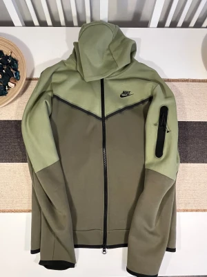 Nike Tech grön hoodiejacka med dragkedja M - Nike hoodiejacka i två nyanser av grönt med svart logga på bröstet. Jackan har hel dragkedja framtill, huva och en praktisk ficka med dragkedja på ärmen. Tillverkad i bomull och polyester för en skön och sportig känsla.Sömmen på ärmen har gått upp, men den är inte förstörd och den kan sys om igen. Unisex. Använd bara 2-3 gånger.
