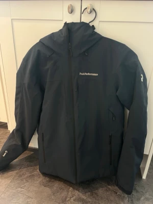 Peak Performance skidjacka  - Snygg blå skidjacka från Peak Performance med hipe som är att den är vattentät. Jackan har en ficka pp bröstet där det finns en duk som man kan torka sina glasögon med.