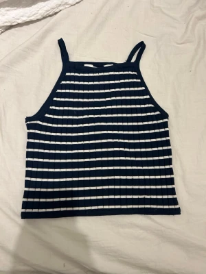 Randig ribbad linnetopp - Snygg ribbad linnetopp med marinblå och vita ränder. Toppen har smala axelband och är ärmlös, perfekt för varma dagar. Materialet känns mjukt och stretchigt, vilket ger en bekväm passform. Den är även ganska kort.