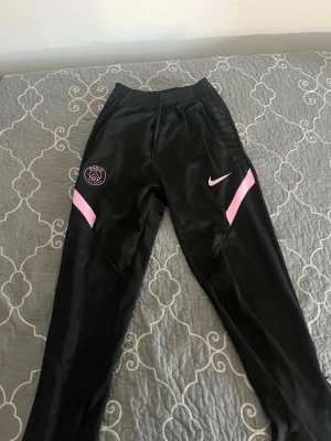 Svarta PSG träningsbyxor Nike - Snygga svarta träningsbyxor från Nike med PSG-logga på vänster ben och rosa detaljer. Byxorna har resår i midjan och är tillverkade i ett glansigt, sportigt material som passar perfekt till träning eller chill. Smal passform och klassisk look.
