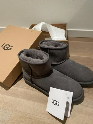 Gråa UGG tofflor - Mysiga gråa tofflor från UGG i mjuk mocka med fluffigt foder. Klassisk rund tå och platt sula, perfekt för att hålla fötterna varma. Snygg och enkel design med UGG-logga på kartongen.