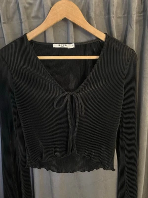 	Elegant svart topp med knyt, plisserad – NA-KD - Svart plisserad topp med knyt – NA-KD Elegant och trendig svart topp i plisserat material från NA-KD. Djup v-ringning med dekorativ knytning framtill och lätt vågig nederkant som ger en feminin silhuett. Figurnära passform och stretchigt tyg som sitter superfint på kroppen.  Perfekt både till fest, middag eller en snygg vardagslook med jeans eller kjol 🖤 Mycket fint skick, sparsamt använd. Storlek: XS  Hör av dig vid frågor eller om du vill ha fler bilder 💌