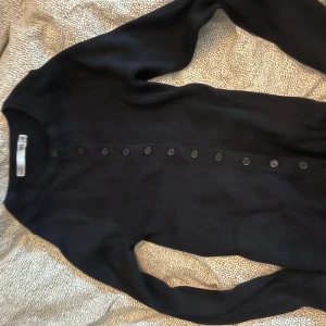 Svart ribbad kofta med knappar - Säljer en svart ribbad kofta med rund hals och långa ärmar. Koftan har knäppning hela vägen framtill med svarta knappar och är i ett mjukt, stretchigt material. Perfekt att styla lager på lager eller bära som den är.