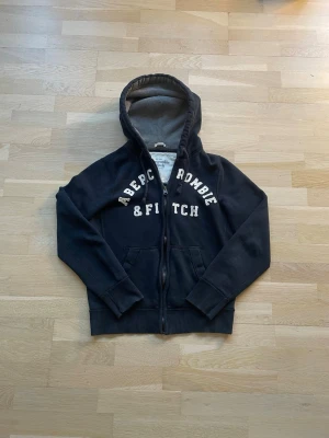 Svart hoodie från Abercrombie & Fitch - Mörkblå hoodie med dragkedja från Abercrombie & Fitch. Stor huva med grått innerfoder, ribbade muddar och fickor framtill. Stora vita bokstäver på bröstet. Perfekt för en avslappnad och cool stil.