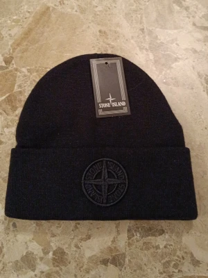 Svart mössa från Stone Island - Snygg svart mössa från Stone Island med uppvikt kant och broderad logotyp framtill. Klassisk ribbstickad design som passar perfekt till streetwear och håller dig varm under kalla dagar. Märket syns tydligt på framsidan för en clean och stilren look.