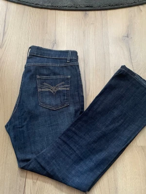 Mörkblå jeans med broderad bakficka - Snygga mörkblå jeans med kontrastsömmar och broderad detalj på bakfickan. Klassisk femficksmodell med bälteshällor och rak passform. Perfekta till en avslappnad stil och tillverkade i mjukt bomullsjeansmaterial.