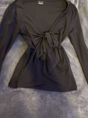 Svart knyttopp - Svart omlottopp från Gina Tricot med lång ärm och snygg knytning framtill. Toppen har en djup v-ringning och är tillverkad i mjukt, stretchigt material som sitter bekvämt på kroppen. Perfekt för att skapa en trendig och stilren look.