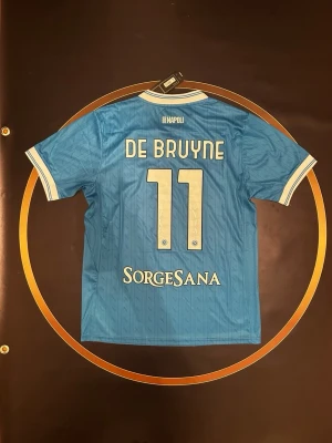 Napoli matchtröja De Bruyne #11 - Ljusblå Napoli matchtröja med De Bruyne och nummer 11 på ryggen. Tröjan har vita detaljer vid krage och ärmslut, samt klubbmärke och sponsorer på framsidan. Tillverkad i lätt och ventilerande material, perfekt för fotbollsträning eller match.