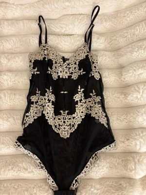 Svart spetsbody från Intimissimi - Snygg svart body från Intimissimi med tunna axelband och vackra spetsdetaljer i beige. Bodyn har blommigt spetsmönster framtill och längs kanterna, samt knäppning i grenen. Perfekt för dig som gillar eleganta underkläder med lyxig känsla.