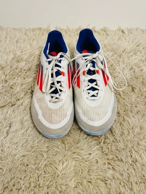 Vita Adidas fotbollsskor med röda detaljer - Säljer ett par vita Adidas fotbollsskor med klassiska röda och svarta ränder på sidorna. Skorna har blå insida, snörning och platt sula. Perfekta för dig som vill ha stilrena och sportiga skor till planen.