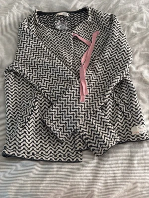 Mönstrad kofta från Odd Molly - Säljer en svartvit stickad kofta från Odd Molly med zigzag-mönster och rosa band framtill. Koftan har långa ärmar och är tillverkad i mjukt material. Perfekt för dig som gillar unika detaljer och snygga mönster.