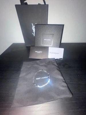Silverarmband från Saint Laurent - Snyggt och stilrent armband från Saint Laurent i silverfärgad metall. Kedjan är tunn och har en liten bricka med logotypen ingraverad. Kommer med originalpåse, ask och certifikat. Perfekt accessoar för dig som gillar exklusiva detaljer.