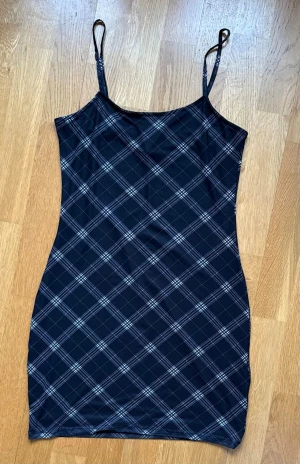 Blå rutig linnetopp med smala band - Snygg blå kortklänning med tunna axelband och rutigt mönster i blått. Klänningen har en tight passform och är tillverkad i ett stretchigt syntetmaterial som sitter skönt på kroppen. Perfekt för dig som gillar en enkel men trendig look.