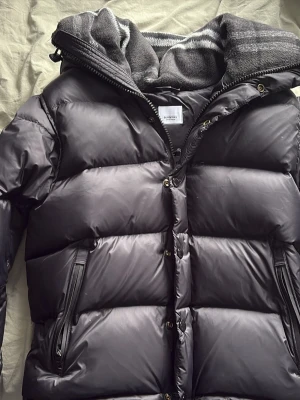 Svart dunjacka från Filippa K - Snygg svart dunjacka från Burberry med dragkedja och knappar framtill. Jackan har huva och flera praktiska fickor med dragkedja. Perfekt för kalla dagar och enkel att styla till olika outfits.