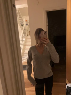 Grå stickad tröja med v-ringning - Mysig grå stickad tröja med v-ringad hals och långa ärmar. Tröjan har en enkel och stilren design, perfekt att matcha med jeans eller kjol. Liten broderad detalj på bröstet. Passar dig som gillar en avslappnad och trendig look.