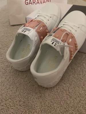 Valentino Rockstud sneakers vit/rosa - Snygga vita sneakers från Valentino med ljusrosa band och ikoniska nitar över vristen. Skorna har rund tå, platt sula och klassisk snörning. Perfekta för dig som vill sticka ut med en lyxig och trendig detalj.  Storlek 39 och är i mycket fint skick. Skorna levereras med original kartong och valentiono påse. Snabb leverans och trygg leverans men ansvarar inte för postens slavar.