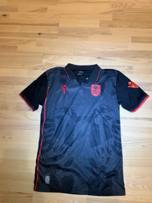 Svart fotbollströja Albanien Macron - Snygg svart fotbollströja från Macron med Albaniens landslagsemblem på bröstet. Tröjan har röda detaljer längs krage, ärmslut och sidor samt diskret mönster i tyget. Perfekt för match eller träning.