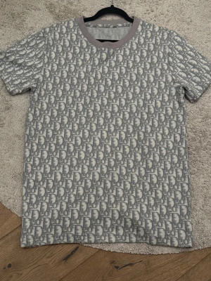 Grått set med Dior-mönster - Snyggt tvådelat set från Dior med t-shirt och shorts i matchande grått och vitt monogrammönster. T-shirten har rund halsringning med ljusgrå kant och shortsen har dragsko i midjan. Perfekt för en trendig och avslappnad look.
