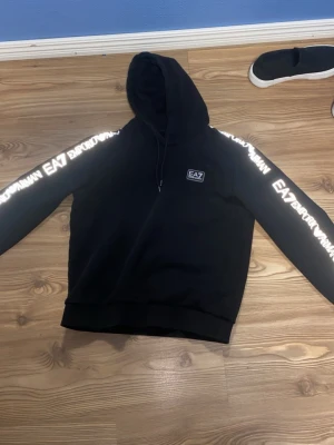 Svart EA7 Emporio Armani set - Snyggt svart set från EA7 Emporio Armani med matchande hoodie och byxor. Setet har vita logotexter längs ärmar och ben samt EA7-märke på bröstet och benet. Hoodien har huva och dragsko, byxorna har resår i midjan. Perfekt för en sportig och trendig look.