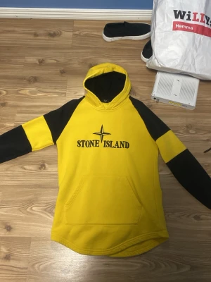 Gul och svart hoodie från Stone Island - Snygg gul hoodie med svarta detaljer från Stone Island. Stor logga framtill, huva och känguruficka. Ärmarna har både gula och svarta partier för en cool kontrast. Perfekt för dig som vill sticka ut med streetwear-stil.