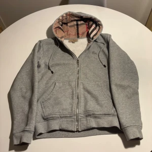 Burrbery zip up tröja  - Grå zip-hoodie/kofta från Burberry i mjuk och bekväm kvalitet. Klassiskt och tidlöst plagg med dragkedja framtill, två fickor och luva med Burberrys ikoniska rutmönster på insidan. Broderad Burberry-logga på bröstet. Ribbstickade muddar vid ärmslut och nederkant.