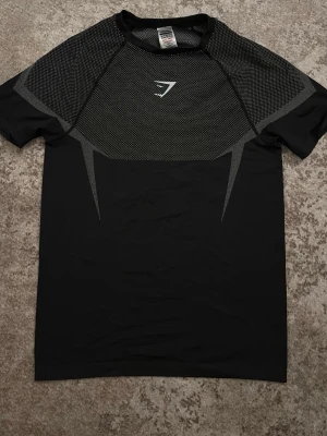 Svart träningströja från Gymshark - Snygg svart träningströja från Gymshark med meshdetaljer över axlar och bröst. Tröjan har en sportig design med logga framtill och är tillverkad i ett lätt, syntetiskt material som andas. Perfekt för gymmet eller löprundan.storlek M/L
