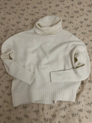 Vit polotröja i stickat material - Klassisk vit stickad polotröja med bred ribbad mudd vid ärmslut och nederkant. Tröjan har en hög, vikt polokrage och är gjord i ett mjukt material som känns skönt mot huden. Perfekt för lager på lager och en stilren look.