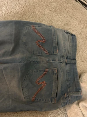 Ljusblå jeans med broderade bakfickor - Säljer ett par ljusblå jeans med unika broderade detaljer i rött på bakfickorna. Jeansen har klassisk femficksdesign, dubbla knappar i midjan och bälteshällor. Perfekta för dig som vill ha något lite extra i vardagen.
