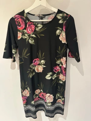 Svart blommig klänning Happy Holly storlek 36/38 - Svart kortklänning från Happy Holly med färgglatt blommönster i rosa och beige toner. Klänningen har rund halsringning, korta ärmar och dekorativt mönster längst ner. Tillverkad i mjukt och stretchigt material för en bekväm passform.