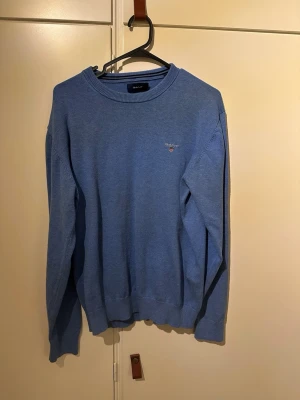 Blå crewneck tröja från Gant - Snygg blå rundhalsad tröja från Gant med diskret broderad logga på bröstet. Tröjan har långa ärmar, ribbade muddar och är gjord i mjukt material som passar perfekt till en avslappnad stil.
