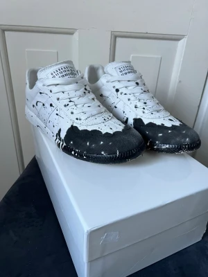 Maison Margiela Gats  svartvita (Äkta) -  (Skriv innan köp)  Unika sneakers från Maison Margiela i vitt tyg med svart färgstänksmönster över tå och sula. Jag säljer skorna eftersom gillade dom inte så mycket. Jag har haft dom i 2 månader nu under vintern vilket man kan se eftersom färgen på tån har blivit lite mot mörk grå då det vart blött ute. Jag kan tänka mig att skorna kommer vara riktigt feta under våren.               Jag köpte skorna för 8799kr från Maison Margiela. Mitt pris är ganska högt då jag inte använt skorna så länge