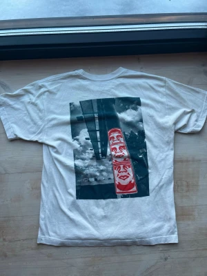 Vit OBEY t-shirt med grafiskt ryggtryck strl L - Vit t-shirt från OBEY med ett stort grafiskt tryck på ryggen som visar en cool fotomontage med skyltar och moln. T-shirten har rund hals och är tillverkad i bomull. Perfekt för dig som gillar streetwear och unika prints.