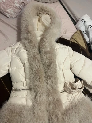 Vit pälsjacka med luva och dragkedja  - Säljer en fluffig vit pälsjacka med stor luva och breda pälsdetaljer längs huva, ärmslut och nederkant. Jackan har dragkedja framtill, två fickor med dragkedja och markerad midja. Perfekt för dig som vill ha en riktigt cozy vinterlook.