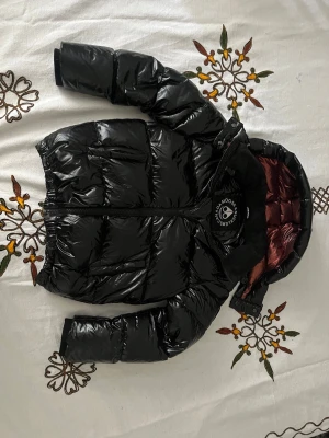 Svart glansig dunjacka från Save The Duck - Snygg svart glansig dunjacka från Save The Duck med huva och dragkedja framtill. Jackan har quiltad design och rödbrunt innerfoder. Perfekt för kalla dagar och ger en modern look.