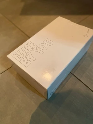 Nike By You vita fotbollsskor - Unika Nike By You fotbollsskor i vitt med blå och lila swoosh-detaljer. Skorna har högt skaft och blå snörning för extra stöd. Tillverkade i syntetmaterial för bästa prestanda på planen. Perfekta för dig som vill sticka ut på matchen.