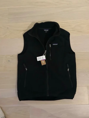 Svart fleeceväst  - Säljer en svart fleecevästen med dragkedja framtill och tre praktiska fickor med dragkedja. Västen har hög krage och är tillverkad i mjuk återvunnen polyester. Perfekt för lager-på-lager och outdoor-vibe.