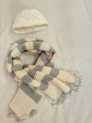 Virkad halsduk i grått och vit och virkad mössa  - Helt nya aldrig använda. Virkade hemma hos mig — bra för hållbarhetsarbete. Mysig virkad mössa med matchande virkad halsduk. Halsduken har en chunky känsla och avslutas med fransar i ändarna. Perfekt för att hålla värmen under kyliga dagar och ger en avslappnad look till din outfit. Längden på halsduken är 119cm.