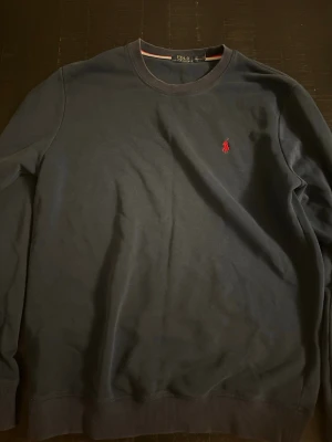 Mörkblå sweatshirt från Polo Ralph Lauren - Klassisk mörkblå sweatshirt från Polo Ralph Lauren med rund halsringning och den ikoniska röda logotypen broderad på bröstet. Tröjan har ribbade muddar vid ärmslut och nederkant. Perfekt för en avslappnad och stilren look.