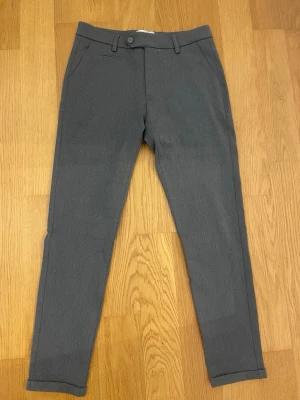 Grå kostymbyxor med smal passform - Snygga grå kostymbyxor med smal, rak passform och klassiska detaljer som bälteshällor och knappstängning. Perfekta för en clean och stilren look. Materialet känns mjukt och har en lätt stretch för extra komfort.