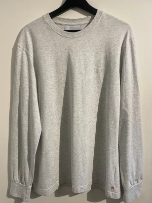 Ljusgrå långärmad t-shirt från Han Kjobenhavn - En stilren ljusgrå långärmad t-shirt med rund halsringning. Tröjan har en enkel och clean design med diskret broderad logga på bröstet och liten detalj nertill. Perfekt för en avslappnad och trendig look.