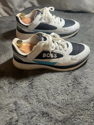 Svarta och vita sneakers från BOSS - Snygga sneakers från BOSS i svart och vitt med blå detalj på sidan. Skorna har rund tå, snörning och en platt sula. Materialet är en mix av syntet och textil, vilket ger en sportig och modern look. Perfekta för dig som gillar stilrena och trendiga sneakers.