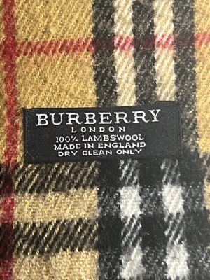 Burberry halsduk i lammull - Klassisk halsduk från Burberry i 100% lammull med det ikoniska rutmönstret i beige, svart, vitt och rött. Halsduken har fransar i ändarna och är tillverkad i England. Perfekt accessoar för att lyfta din outfit.