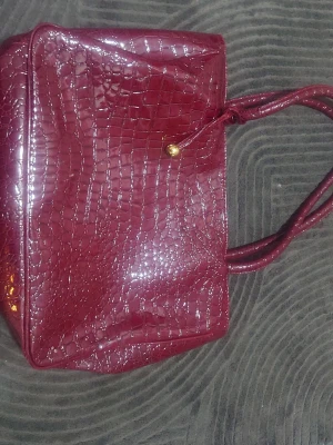 Vinröd handväska i krokopräglat skinn - Snygg vinröd handväska med krokopräglat mönster och glansig finish. Väskan har dubbla handtag och stängs med en guldfärgad knapp. Insidan är fodrad i matchande färg och har praktiska innerfack för småsaker.