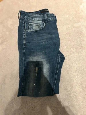 Blå jeans från Alessandro Zavetti - Snygga blå jeans från Alessandro Zavetti med slitna detaljer och diskreta vita stänk på både fram- och baksida. Jeansen har klassisk femficksmodell, normal passform och är tillverkade i mjukt denimtyg. Perfekta för en avslappnad och trendig look.